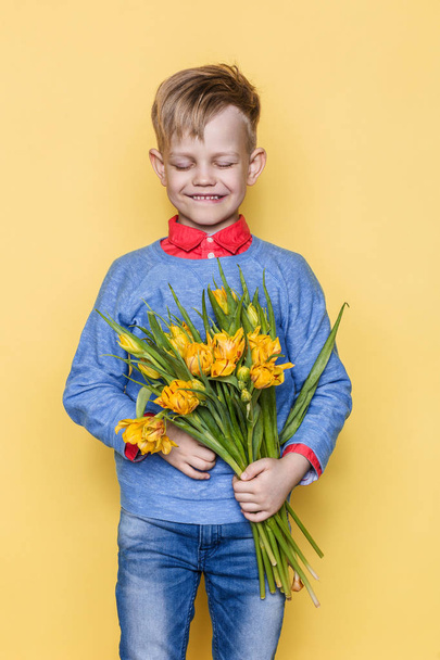 Little beautiful child with a bouquet of tulips. Son gives mom flowers on Women's Day, Mother's Day. Birthday. Valentine's day. Spring. Studio portrait over yellow background - Photo, image