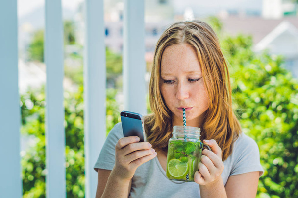 A young woman holds a mason jar in her hand with a mojito and a smartphone. Spring drink concept - Photo, Image