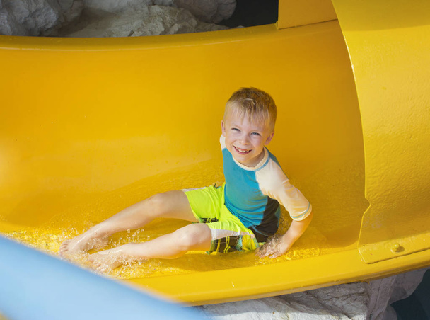 Lächelnder kleiner Junge reitet eine gelbe Wasserrutsche hinunter - Foto, Bild