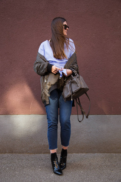 perfect spring fashion outfit. european fashion blogger wearing a trendy crossover striped blouse with a front knot, a bomber jacket, jeans, ankle boots and holding a stylish handbag. - Photo, Image