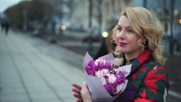 Young blonde woman with flowers bouquet on a city street - Felvétel, videó