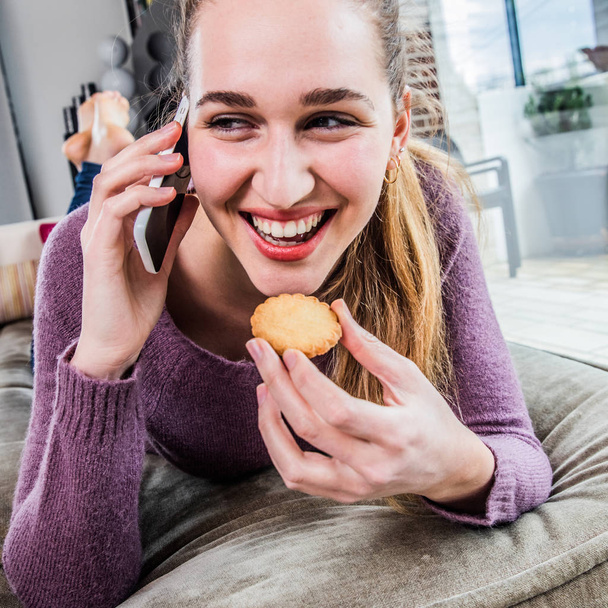 laughing gorgeous young woman talking on smartphone, eating a cookie - Foto, afbeelding