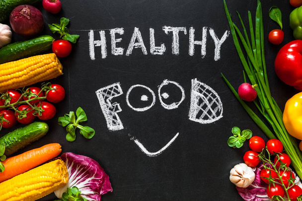 Photo of a table top full of fresh vegetables or healthy food background. Healthy food concept with fresh vegetables for cooking.Title "Healthy food" with smile is written by chalk on the middle of dark background. - Foto, imagen