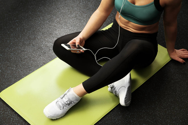 Jovem com telefone inteligente e fones de ouvido sentados no tapete em um ginásio
 - Foto, Imagem
