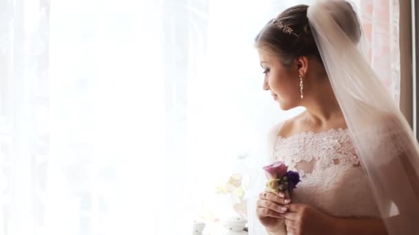 Mooie bruid in luxe trouwjurk op zoek door het raam voordat de ceremonie van het huwelijk. Vrouw bedrijf bloemen - Video