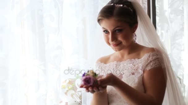Krásná nevěsta v luxusních svatebních šatech u okna před svatební obřad. Žena hospodářství květiny - Záběry, video