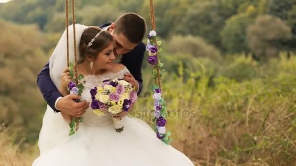 Groom kissing and swinging his beautiful bride in white wedding dress with flower bouquet on swing in summer forest, park - Footage, Video