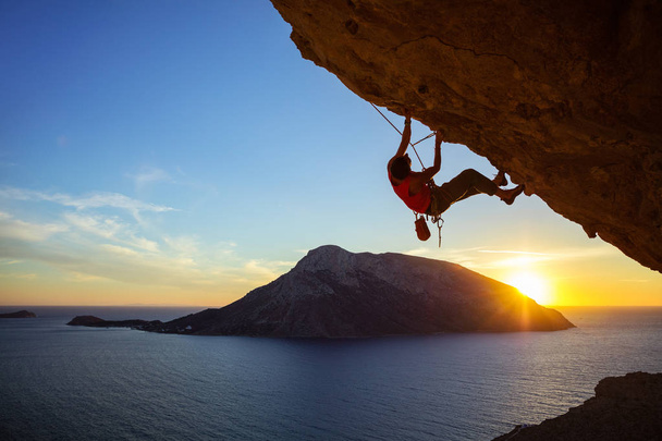 若い男が日没時突出の崖を登る - 写真・画像