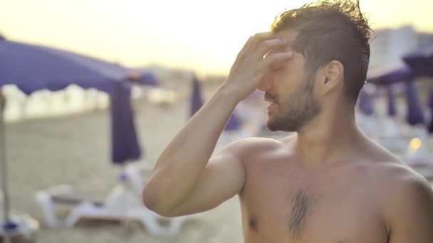かわいい若い男に触れる夕暮れ背景の砂と太陽のベッドでビーチで彼の髪立っています。スローモーション - 映像、動画