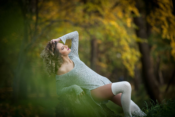 sensual girl with long legs sitting on a stump in an autumnal scene.Long legs attractive blonde with curly hair relaxing in autumnal park.Fashionable young woman posing on a stump in the forest. - 写真・画像