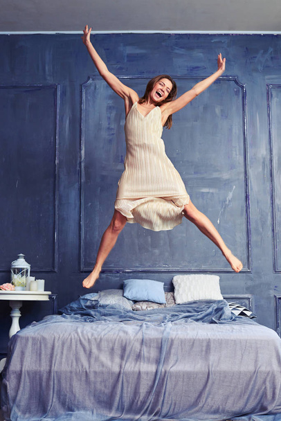 Extremely excited woman in nightgown jumping on bed with arms an - 写真・画像
