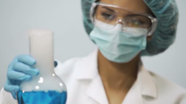 Female chemist holding flask with blue evaporating liquid in hand, experiment - Imágenes, Vídeo