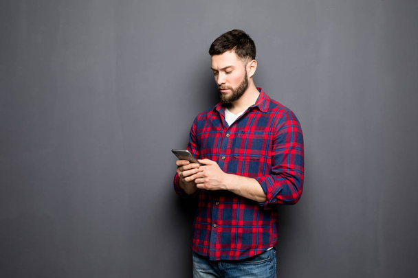 Уверенный молодой человек держит смартфон и смотрит на него, стоя на сером фоне
 - Фото, изображение