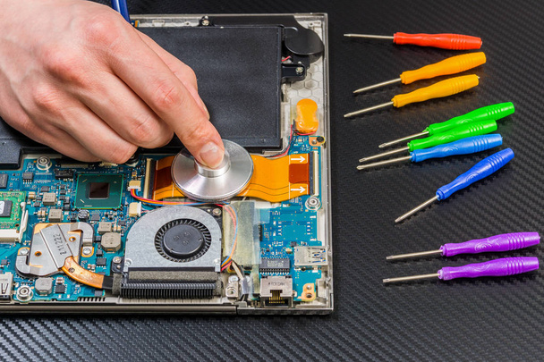 person hands diagnostic laptop mainboard with stethoscope on carbon background with rainbow collection of screwdrivers - Photo, Image
