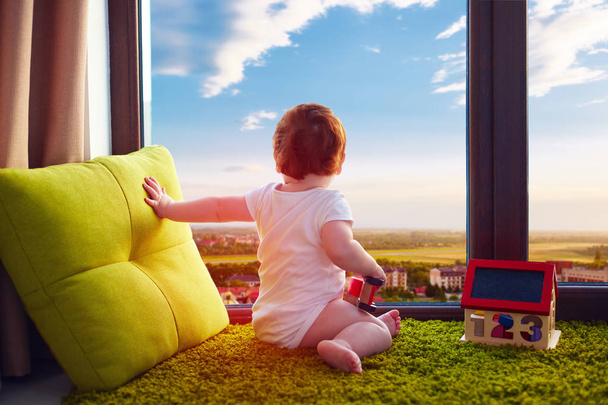 infant baby sitting on carpet at home and watches the beautiful cityscape through the window - Foto, Imagem
