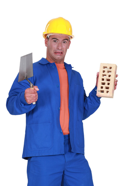 Mason holding a trowel and a brick and looking disgusted - Фото, зображення