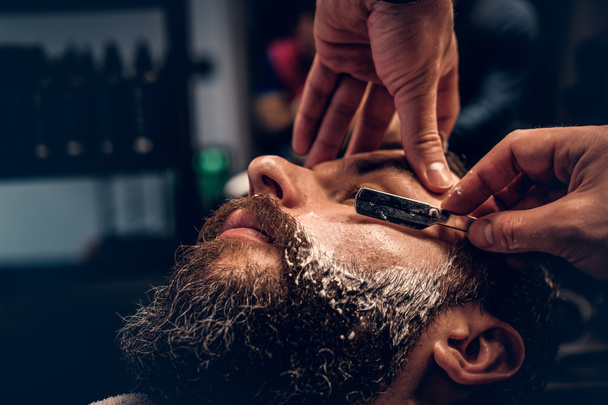 Barber shaving bearded male with a sharp razor - Photo, Image