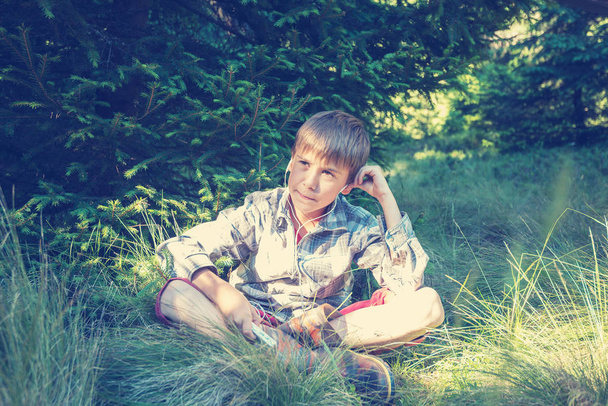 Sonhando menino em fones de ouvido senta-se na grama exuberante
  - Foto, Imagem