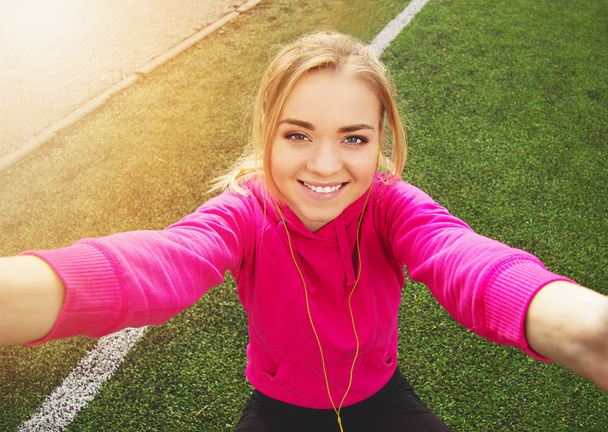 Young beautiful blonde hair fitness girl doing selfie on a camera on the stadium. Summer sport activity. - Фото, зображення