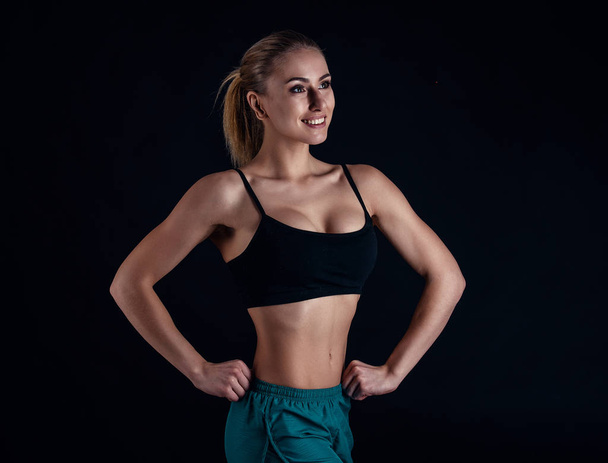 Sporty young girl in sportswear showing muscles on black background. Tanned young athletic woman. A great sport female body. - Фото, изображение