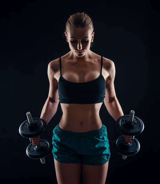Portrait of a young fitness woman in sportswear doing workout with dumbbells on black background. Tanned sexy athletic girl. A great sporty female body. - Фото, зображення