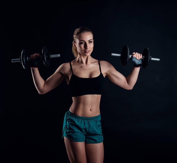 Cute athletic model girl in sportswear with dumbbells in studio against black background. Ideal female sports figure. Fitness woman with perfect sculpted muscular and tight body. - 写真・画像