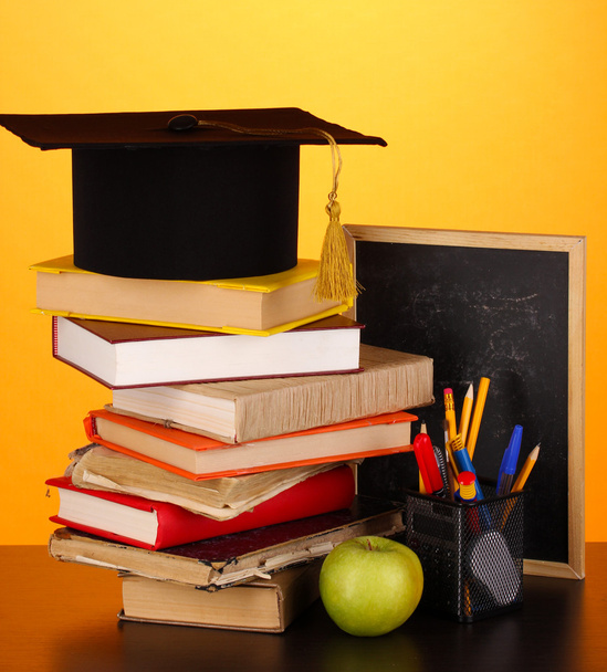 Books and magister cap against school board on wooden table on yellow background - Fotografie, Obrázek