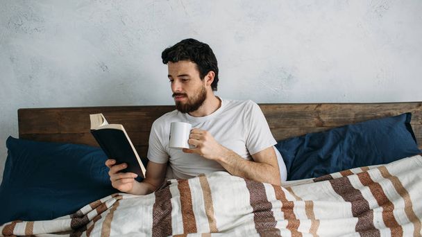 Bearded man reading a big book lying in his bedroom. - Valokuva, kuva