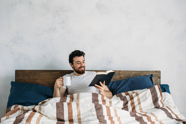 Bearded man reading a big book lying in his bedroom. - 写真・画像