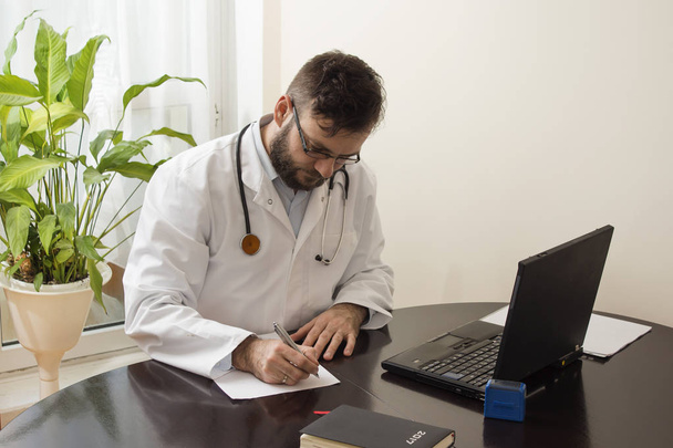 Ein Arzt im weißen Kittel in einer Arztpraxis sitzt an einem Tisch und füllt Papiere. ein Arzt bei der Arbeit.  - Foto, Bild