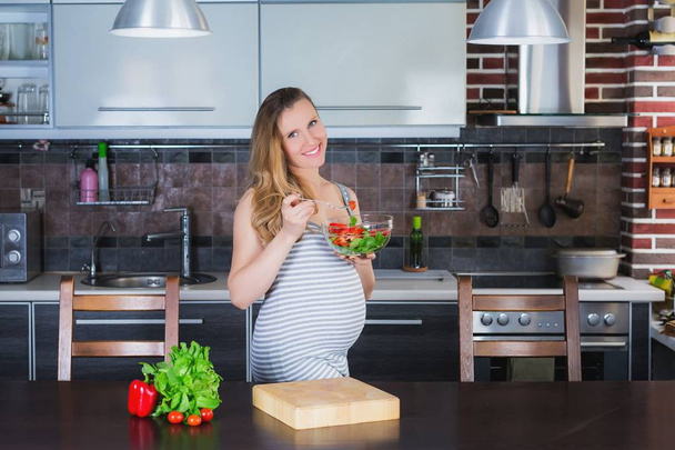 The smiling pregnant woman in kitchen is eating vegetable salad. - Fotoğraf, Görsel