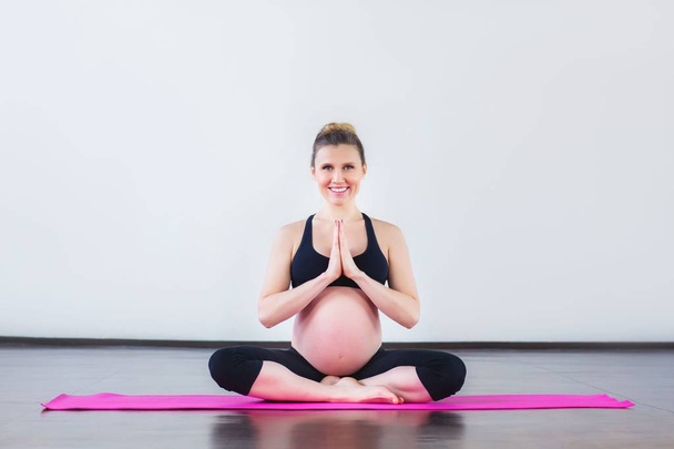 Young pregnant woman sitting in lotus position in gym. Pregnancy Yoga and Fitness. - 写真・画像
