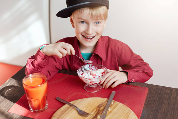 A happy young boy with blue eyes and blond hair dressed in red shirt and stylish cap siting at cosy caffe eating delicious ice-cream and drinking tomato juice looking happily into a camera - Fotoğraf, Görsel