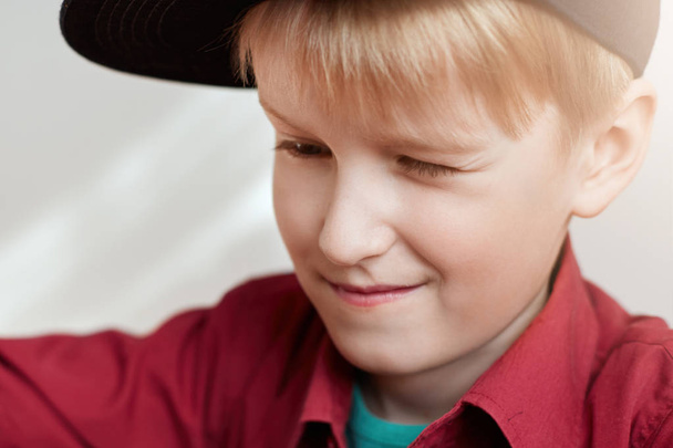 A close-up of curious little boy with white hair wearing stylish black hat and red shirt looking at something with narrowed eyes. A horizontal portrait of cute child exploring something - Foto, Bild