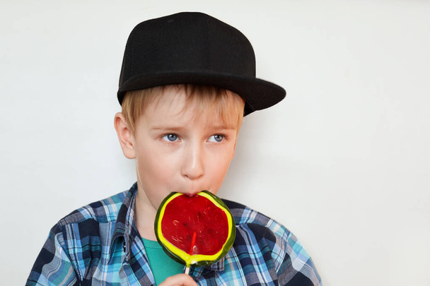A portrait of a cute little blond boy in black cap and checked shirt eating a bright lollipop isolated over white background looking into distance. A handsome boy with blue eyes licking candy - Fotoğraf, Görsel