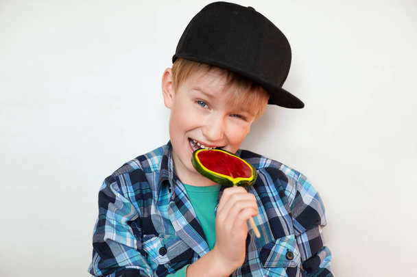 Headshot of beautiful little boy holding and biting lollipop looking at the camera with happy and joyful expression. Handsome boy in stylish clothes isolated over white background with sweet candy  - Фото, изображение