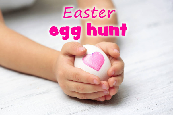 Little child holding egg on table. Text EASTER EGG HUNT on background - Photo, image