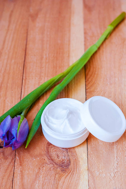 Crema facial en un frasco blanco sobre un fondo de madera rugosa con un tulipán morado
. - Foto, imagen