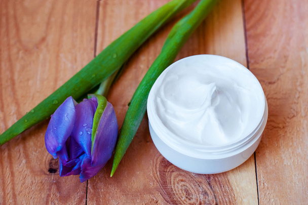 Crema facial en un frasco blanco sobre un fondo de madera rugosa con un tulipán morado
. - Foto, Imagen