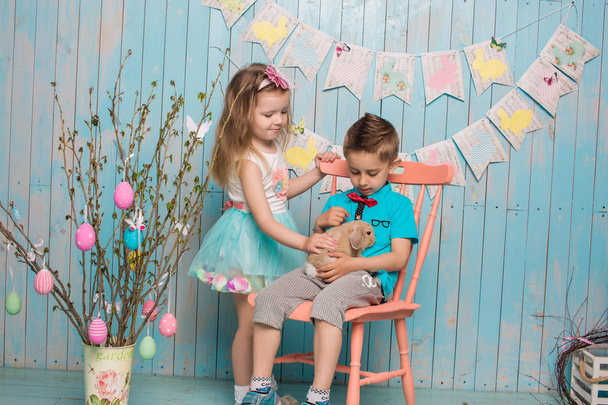 Dos niños pequeños, niño, niña, hermano y hermana junto con el conejo sentado en el suelo en ropa azul brillante Pascua, huevos, humor festivo, emoción y sonrisa fiesta sorpresa celebración
 - Foto, imagen