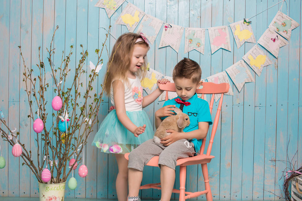 Dos niños pequeños, niño, niña, hermano y hermana junto con el conejo sentado en el suelo en ropa azul brillante Pascua, huevos, humor festivo, emoción y sonrisa fiesta sorpresa celebración
 - Foto, imagen