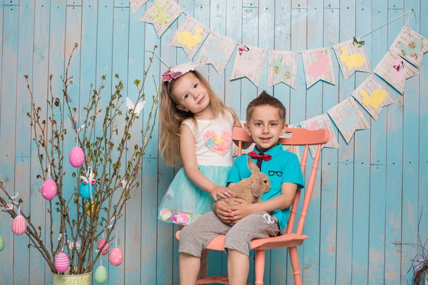 zwei kleine Kinder, Junge, Mädchen, Bruder und Schwester zusammen mit Kaninchen auf dem Boden sitzend in leuchtend blauer Kleidung Ostern, Eier, festliche Stimmung, Emotionen und Lächeln Überraschung Feiertagsfeier - Foto, Bild