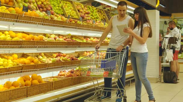 Mladý pár kavkazské chůzi v supermarketu s vozíkem trhu a volbou čerstvá jablka. Člověk je zprovoznění obchod košík ovoce. Žena používá počítač tablet pc zkontrolovat nákupní seznam - Záběry, video