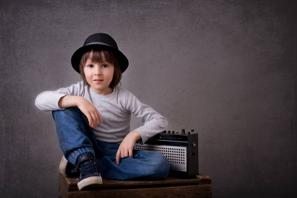 Cute boy with black hat, sitting on a wooden tray, holding radio - Фото, изображение