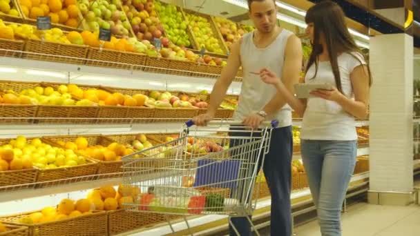 Joven pareja caucásica caminando en un supermercado con un carrito de mercado y la elección de manzanas frescas. El hombre está poniendo frutas en la cesta de la tienda. Mujer está utilizando la tableta PC para comprobar la lista de compras
 - Metraje, vídeo