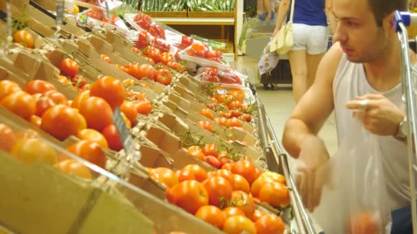 Ein junger Mann wählt im Supermarkt Tomaten und lächelt. glücklicher Kerl, der frische reife rote Tomaten im Lebensmittelgeschäft auswählt, produziert Abteilung. - Filmmaterial, Video