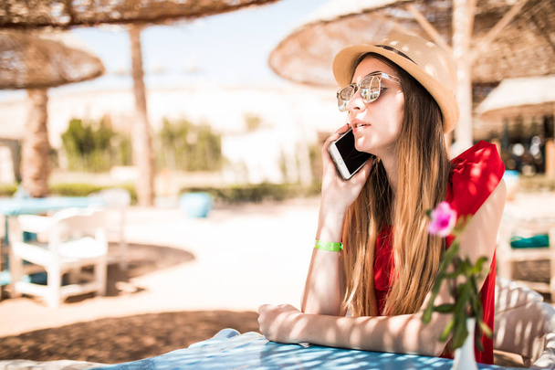 Young woman waiting for order and speak on phone in the restaurant near the sea. Summer vocation - Фото, изображение