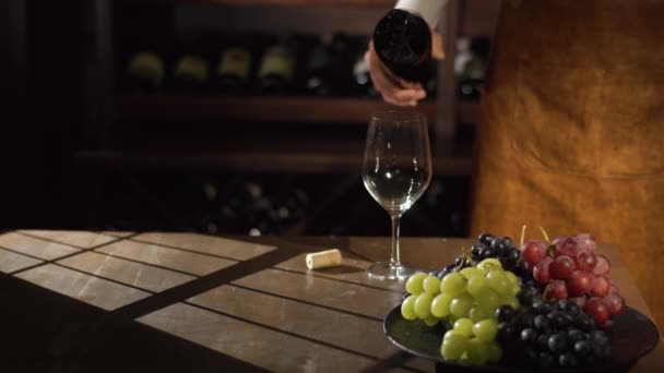 Detail přední portrét sommelier nalil bílé víno z láhve do skla poblíž talíř plný barevných hroznů. Sluneční světlo na stůl. - Záběry, video
