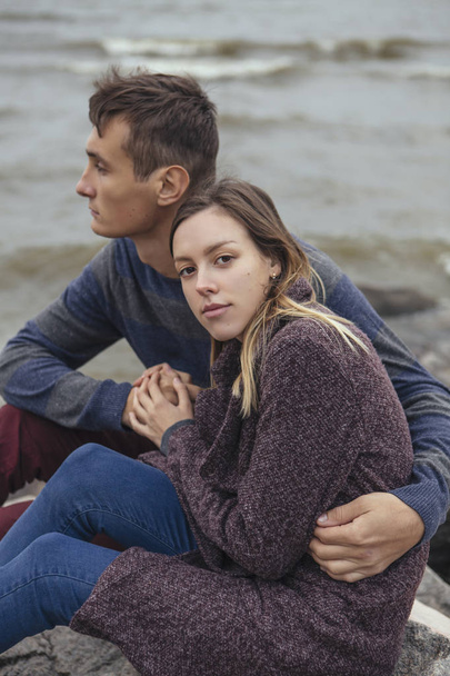 Feliz pareja pensativa sentada en una playa de rocas cerca de abrazos al mar
 - Foto, imagen