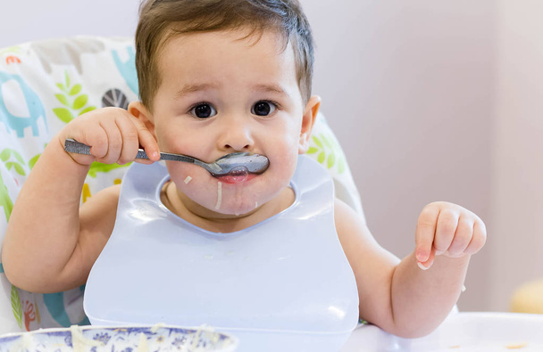 Hymyilevä lapsi syö ruokaa keittiössä. 1-vuotias vauva syö omaa keittoaan. ensimmäinen kiinteät elintarvikkeet, miten opettaa lapselle syödä lusikalla. söpö vauva syö lusikalla lounaaksi
 - Valokuva, kuva